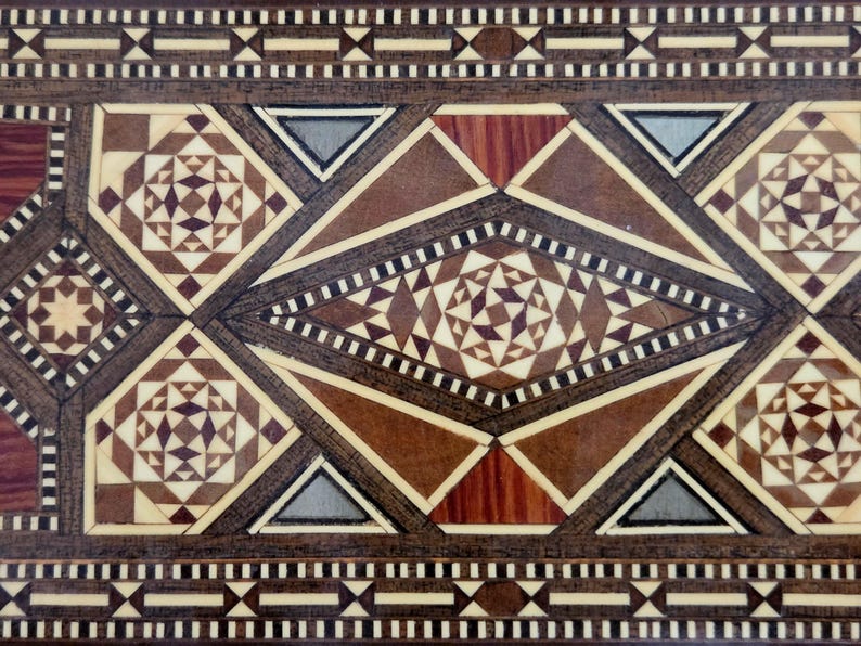 May include: Close-up of a wooden surface with intricate geometric patterns. The design features various shapes, including diamonds, triangles, and stars, in shades of brown, beige, and gray. The detailed craftsmanship suggests a decorative or ornamental item.