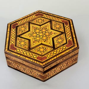 Puede incluir: Una caja hexagonal de madera con intrincados incrustaciones de mosaico. La tapa presenta un diseño de estrella en tonos amarillos, marrones y naranjas. Los lados de la caja están decorados con patrones geométricos. La caja es probablemente para joyas o pequeños recuerdos.