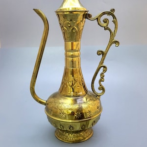 Puede incluir: Una jarra de metal de color dorado con un diseño ornamentado y un largo pico. La jarra tiene un asa con un diseño decorativo de rollo. La base de la jarra tiene la palabra "Jerusalén" inscrita en ella.