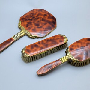 Könnte beinhalten: Ein Vintage-Schminkset aus Schildpatt mit einem Handspiegel, einer Haarbürste und einer Kleiderbürste. Das Set verfügt über goldfarbene Akzente und ein Schildpattmuster.