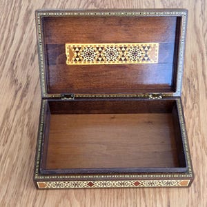 May include: Open wooden box with intricate inlaid geometric patterns in shades of brown, beige, and red. The box is rectangular with a hinged lid and a decorative border. The interior is empty, revealing the natural wood grain.