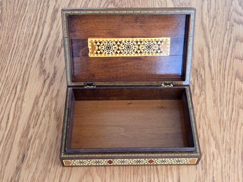 May include: Open wooden box with intricate inlaid geometric patterns in shades of brown, beige, and red. The box is rectangular with a hinged lid and a decorative border. The interior is empty, revealing the natural wood grain.