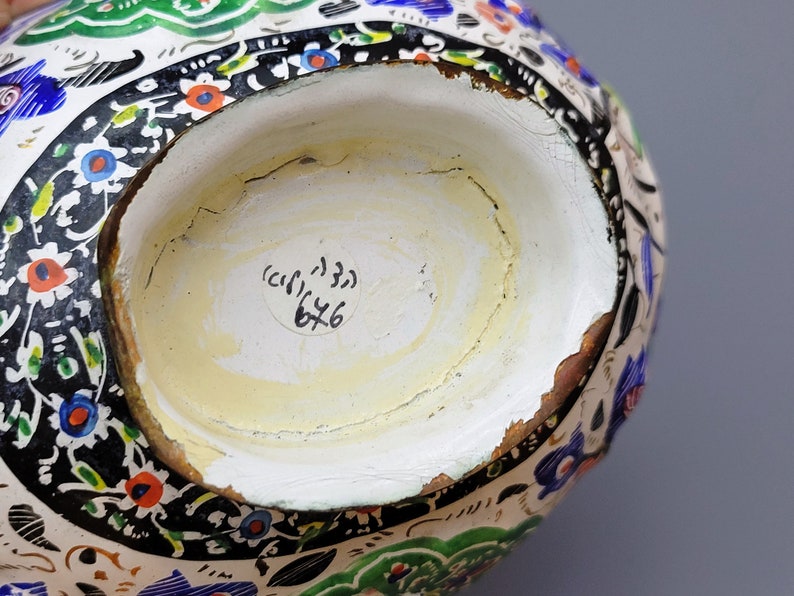 May include: The bottom of a white ceramic pitcher with a black, green, and blue floral design. A white sticker with the text "(c) 133 6x6" is on the bottom.