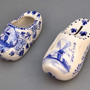 Puede incluir: Dos zuecos de cerámica, pintados en azul y blanco, con diseños de molinos de viento y florales. Un zueco tiene una ranura en la parte superior, lo que sugiere que es un banco. El otro zueco está abierto. La palabra "Holland" está escrita en un zueco.
