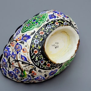 May include: A white ceramic bowl with a black, blue, green, and orange floral design. The bowl has a scalloped edge and a white interior with a handwritten inscription in black.