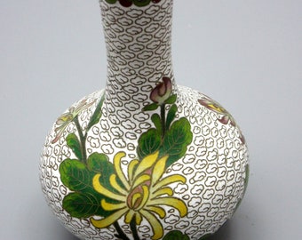 May include: A white cloisonné vase with a narrow neck and a round body. The vase is decorated with green leaves, yellow flowers, and pink and white blossoms. The background is a white pattern of small circles.