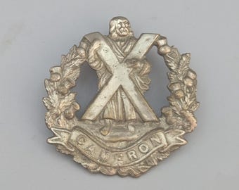 May include: A silver-toned metal military cap badge featuring a central figure standing before a large saltire cross. The figure is encircled by a wreath of thistle leaves and flowers, with a banner at the base bearing the text CAMERON.