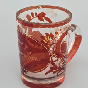 May include: A clear glass mug with a handle, decorated with a red and gold floral pattern. The mug has a red rim and a red band with faded text. The design includes leaves and floral patterns.