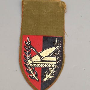 Puede incluir: Un parche militar vintage con un diseño de escudo. El escudo está dividido en mitades rojas y negras, con una ilustración de tanque y ramas de laurel. El parche tiene un respaldo de tela verde y una parte superior de color canela.