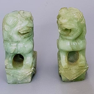Może przedstawiać: Para zielonych jadeitowych figurek foo dog. Figurki są rzeźbione w tradycyjnym chińskim stylu i mają skomplikowane detale. Obie stoją na kwadratowych podstawach.