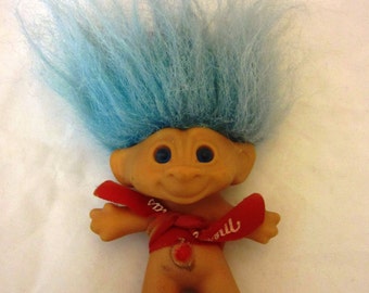 May include: A vintage troll doll with blue hair, a smiling face, and a red ribbon around its waist. The doll has blue eyes and a small, round belly button. The ribbon has white text on it.
