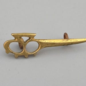 Puede incluir: Pequeño objeto metálico dorado con un diseño único. Cuenta con dos orificios para los dedos conectados a una hoja puntiaguda y una pequeña protuberancia cilíndrica marrón. El objeto tiene una apariencia vintage o antigua.