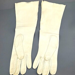 May include: A pair of off-white, long leather gloves. The gloves are smooth and have a classic design, with a slightly textured appearance. The gloves are laid out on a gray surface.