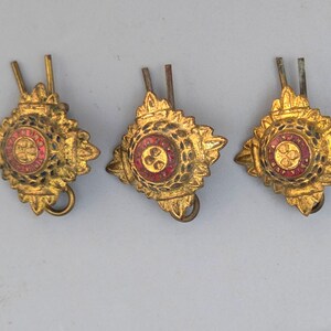 Op de afbeelding: Drie antieke goudkleurige metalen badges met een rode kern en ingewikkelde details. Elke badge heeft een ruitvormig frame met een cirkelvormig middendesign. De badges hebben een speld op de achterkant.