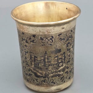 Puede incluir: Una taza metálica con acabado dorado que presenta una escena de paisaje grabada en oscuro con edificios y árboles dentro de un borde ornamental. El recipiente cilíndrico tiene un borde liso y ensanchado y una base sencilla, con el número 84 visible.