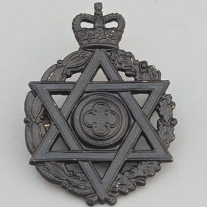 Könnte beinhalten: Ein schwarzes Metallabzeichen mit einem Davidstern in einem Kranz, gekrönt von einer Krone. Die Mitte des Sterns hat ein kreisförmiges Design mit einer kreuzartigen Form. Das Abzeichen hat ein Vintage-Aussehen.
