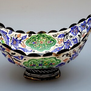 May include: A white ceramic bowl with a scalloped rim and a black base. The bowl is decorated with a colorful floral pattern in shades of blue, green, and gold.