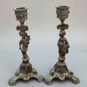 Puede incluir: Un par de candelabros antiguos de color plateado. Cada candelabro presenta una base detallada, una figura central y una parte superior en forma de copa. Las figuras parecen ser un niño y una niña. Las bases tienen un diseño ornamental y enrollado.