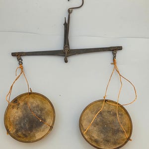 Puede incluir: Una balanza antigua con un marco de metal oscuro y dos cuencos redondos de madera suspendidos por hilo. La balanza tiene un gancho curvo en la parte superior para colgar.