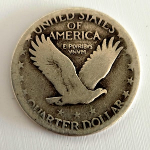 Op de afbeelding: Een zilveren Quarter Dollar munt uit de Verenigde Staten. De munt toont een adelaar met uitgespreide vleugels, omringd door de woorden "UNITED STATES OF AMERICA" en "QUARTER DOLLAR". De munt heeft ook de inscriptie "E PLURIBUS UNUM".