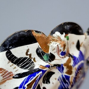 May include: Close-up of a ceramic bowl with a white background and a black, blue, and gold floral design. The bowl has a scalloped edge and shows signs of wear and tear.