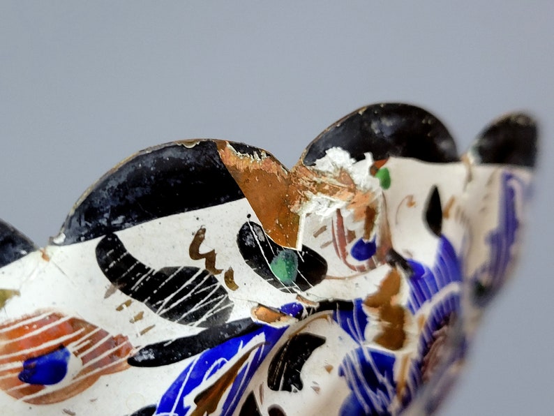 May include: Close-up of a ceramic bowl with a white background and a black, blue, and gold floral design. The bowl has a scalloped edge and shows signs of wear and tear.