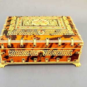 May include: An antique rectangular jewelry box with a tortoiseshell pattern. The box features intricate ivory-colored accents, decorative openwork, and a detailed floral design on the lid. The box is supported by small, ornate feet.