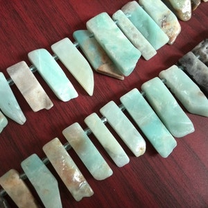 Puede incluir: Una hebra de cuentas de piedras preciosas de color verde claro y marrón, cada cuenta tiene una forma rectangular larga y plana con un borde ligeramente redondeado. Las cuentas están enhebradas en un hilo transparente.
