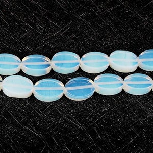 May include: Two strands of oval-shaped, opalescent beads with a white and blue color scheme. Each bead has a subtle, striped design. The beads are strung together, creating a necklace or bracelet component. The background is a textured black surface.