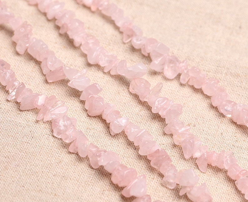 May include: A strand of pink rose quartz chips, also known as rose quartz nuggets, is shown in a close-up image. The chips are irregularly shaped and have a natural, unpolished finish.