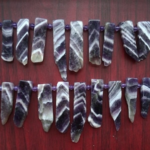 May include: Two strands of amethyst gemstone beads, each strand is composed of multiple purple and white striped, faceted, teardrop-shaped beads. The beads are strung on a clear elastic cord.