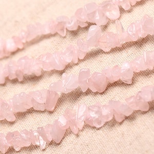 May include: A strand of pink rose quartz chips, also known as rose quartz nuggets, on a light beige fabric background.