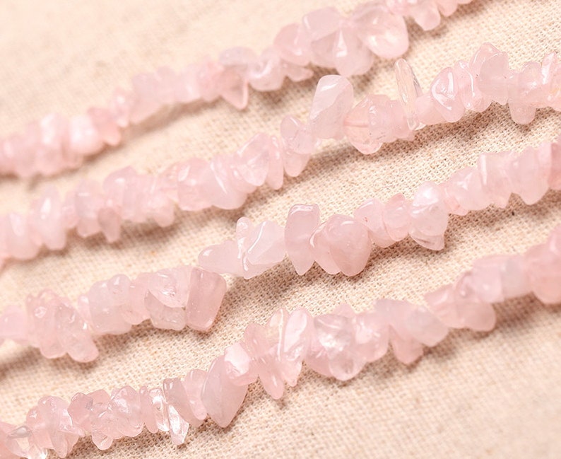 May include: A strand of pink rose quartz chips, also known as rose quartz nuggets, on a light beige fabric background.