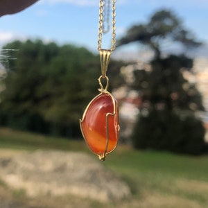K&ouml;nnte beinhalten: Eine goldene Kettenkette mit einem Anh&auml;nger, der einen gro&szlig;en, orangefarbenen Karneol-Stein zeigt, der in Golddraht gewickelt ist.