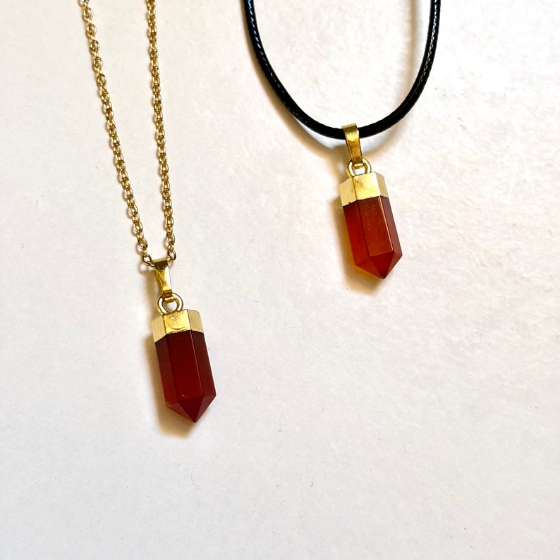 Pendentifs en pierre rouge taillés en pointe, sertis d’un capuchon doré et accroché à une chaîne délicate ou à un cordon noir. Accessoire intemporel et raffiné