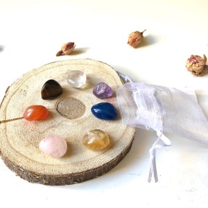 Peut inclure: Un ensemble de sept pierres de chakra roulées sur une tranche de bois, avec un sac en organza blanc. Les pierres sont du quartz clair, de l'œil de tigre, de la cornaline, du quartz rose, de la citrine, de l'améthyste et du lapis-lazuli.