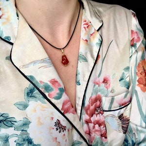 K&ouml;nnte beinhalten: Eine goldene Halskette mit einem roten Karneol-Anh&auml;nger. Die Kette tr&auml;gt eine Person, die ein wei&szlig;es, blumiges Hemd tr&auml;gt.