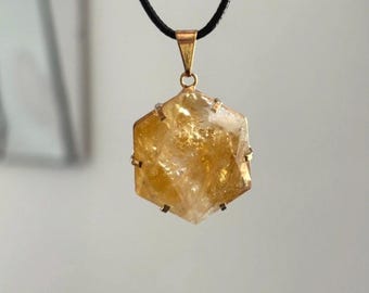 Puede incluir: Un colgante hexagonal de citrino con una fianza y un engaste dorados, suspendido de un cordón negro. La piedra de citrino tiene un tono amarillo dorado translúcido. El colgante es una joya.