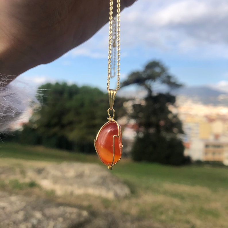 K&ouml;nnte beinhalten: Eine goldene Kettenkette mit einem Anh&auml;nger, der einen gro&szlig;en, orangefarbenen Karneol-Stein zeigt, der in Golddraht gewickelt ist.