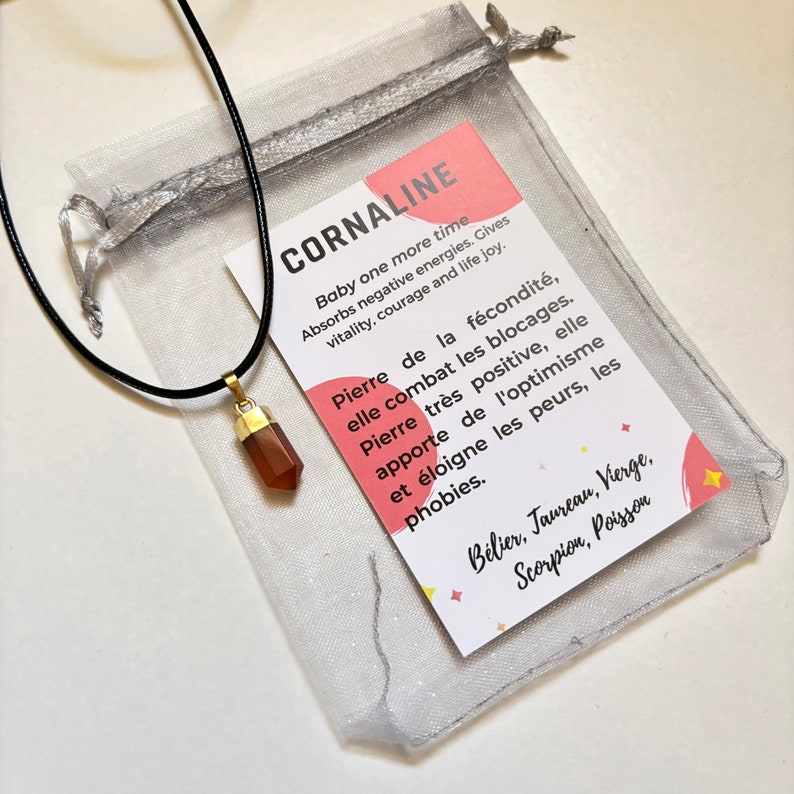 Pendentif en pierre rouge  taillé en pointe, serti d’un capuchon doré et accroché à un cordon noir, doté d’une fiche expliquant les vertus de la pierre et d’une petite pochette