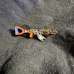 May include: A black metal pin of an orange and blue assault rifle with a black background.