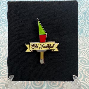 May include: A yellow and brown enamel pin with a red, green, and yellow geyser and a banner that reads "Old Faithful".