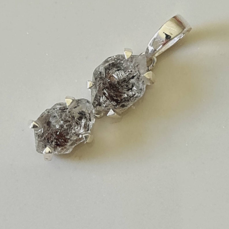 May include: A silver pendant featuring two raw, clear quartz crystals with a rough, faceted texture. The crystals are attached to a silver bail with a loop for attaching a chain.