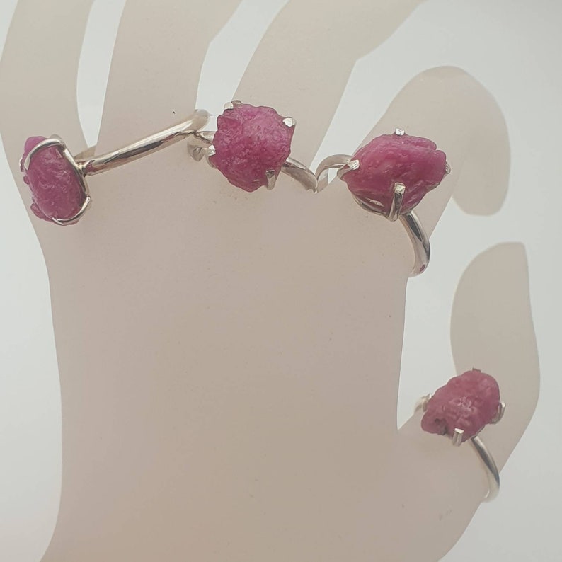 Peut inclure: Quatre bagues en argent avec des pierres pr&eacute;cieuses de rubis brut roses. Les bagues sont pr&eacute;sent&eacute;es sur un mannequin de main blanc.