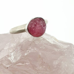 Op de afbeelding: Een zilveren ring met een grote, ruwe, roze edelsteen. De edelsteen heeft een ruw, getextureerd oppervlak en is gezet in een eenvoudige zilveren band. De ring wordt gepresenteerd op een lichtroze, getextureerd oppervlak.