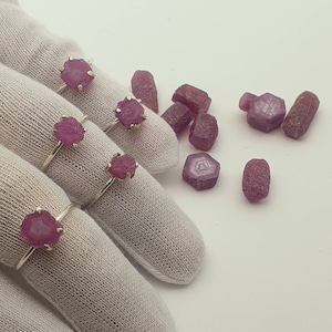 May include: Four silver rings with pink ruby gemstones set in the centre of each ring. The rings are displayed on a white fabric surface. There are several loose pink ruby gemstones scattered around the rings.