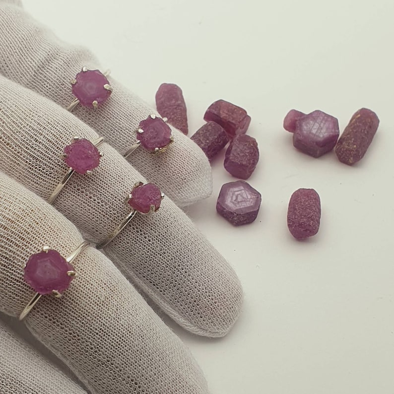 May include: Four silver rings with pink ruby gemstones set in the center of each ring. The rings are displayed on a white fabric surface. There are several loose pink ruby gemstones scattered around the rings.