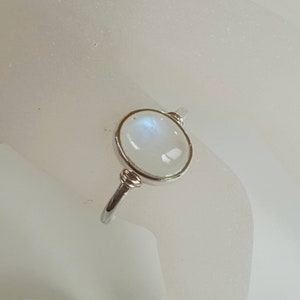 May include: A silver ring with an oval moonstone. The moonstone is milky white with a blue sheen. The ring has a thin, polished band and decorative features near the stone. The ring is a piece of jewellery.
