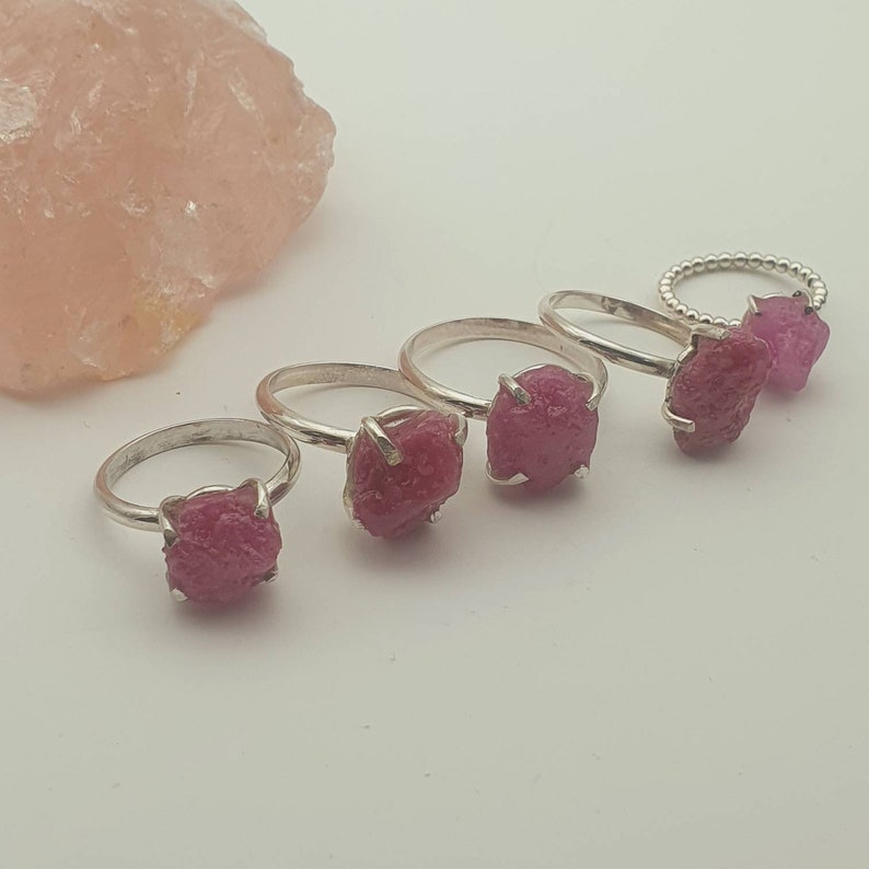 Peut inclure: Cinq bagues en argent avec des pierres pr&eacute;cieuses de rubis brut roses. Les bagues sont dispos&eacute;es en rang&eacute;e sur une surface blanche.