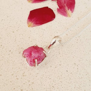 May include: A silver chain necklace with a pendant featuring a raw pink ruby gemstone. The ruby is set in a silver frame and has a rough, natural appearance.